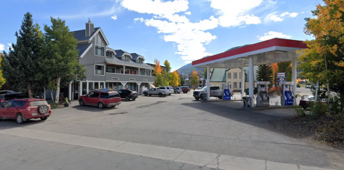 A gas station next to a multi-story building, surrounded by parked cars and autumn trees under a blue sky.