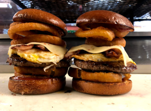 Two towering burgers with beef patties, melted cheese, fried eggs, and onion rings, stacked on toasted buns.