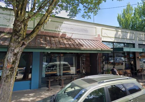 A street view of a storefront with a metal roof, large windows, and outdoor seating under a tree.