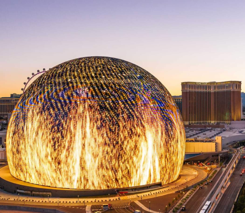 A large, illuminated dome structure with vibrant patterns, set against a sunset sky in a cityscape.