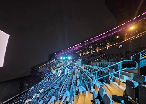A wide view of an empty stadium at night, featuring rows of seats and colorful stage lights in the background.