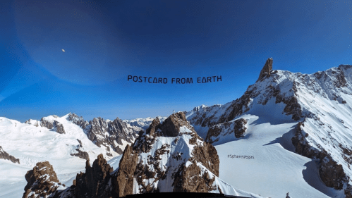 A breathtaking mountain landscape with snow-covered peaks under a clear blue sky, captioned "Postcard from Earth."