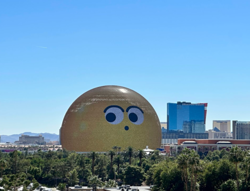 A large, round building with a cartoonish face stands out against a clear blue sky and city skyline.