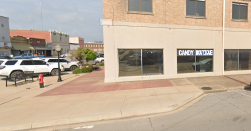 A street view of a candy boutique with large windows, surrounded by parked cars and a small plaza area.