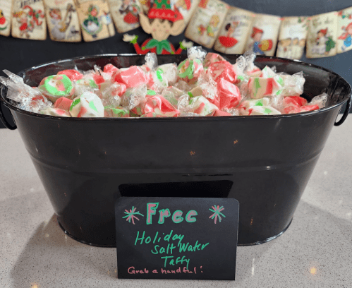 A black bucket filled with red and green holiday saltwater taffy, with a sign that says "Free Holiday Salt Water Taffy!"