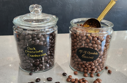 Two glass jars filled with dark chocolate cranberries and milk chocolate raisins, with a golden spoon in one jar.