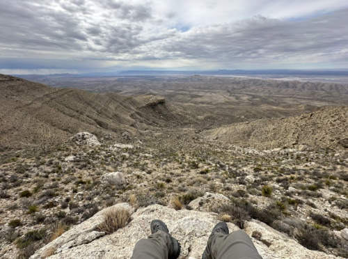 A person sits on a rocky ledge, overlooking a vast, arid landscape under a cloudy sky.