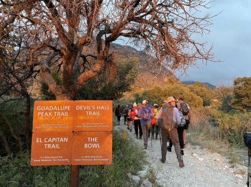 Hikers on a trail with signs for Guadalupe Peak, Devil's Hall, El Capitan, and The Bowl at sunset.