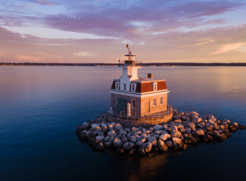 A lighthouse on a rocky island at sunset, surrounded by calm waters and a colorful sky.