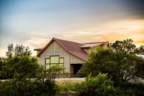 A modern wooden house with a metal roof, surrounded by greenery, under a colorful sunset sky.
