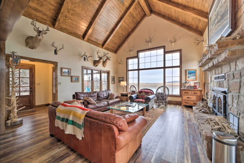 Cozy living room with wooden beams, leather sofas, a stone fireplace, and large windows overlooking a scenic view.