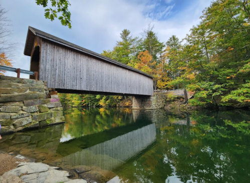 A wooden covered bridge spans a calm river, surrounded by colorful autumn trees and a clear blue sky.