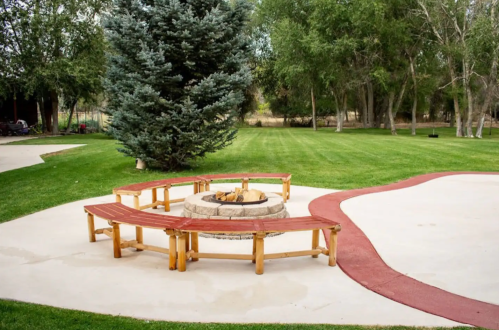 A circular seating area with wooden benches surrounds a stone fire pit on a grassy lawn. Trees in the background.