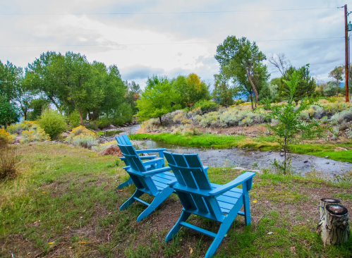 Two blue chairs facing a serene stream, surrounded by lush greenery and trees under a cloudy sky.