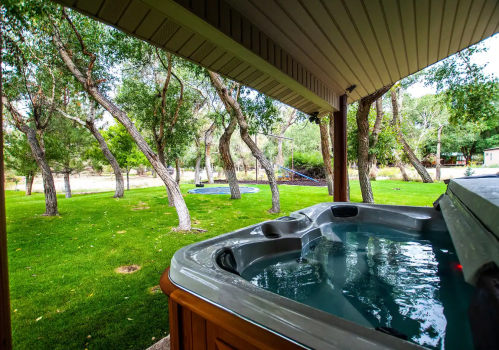 A hot tub on a patio overlooking a lush green yard with trees and a distant play area.