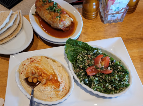 A plate featuring hummus with pine nuts, tabbouleh salad, and a stuffed grape leaf, with pita bread in the background.
