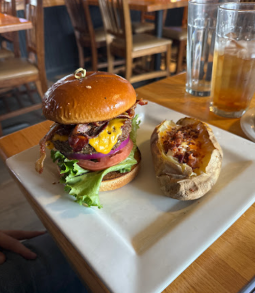 A juicy burger with bacon, cheese, lettuce, and tomato on a plate, alongside a baked potato with toppings.