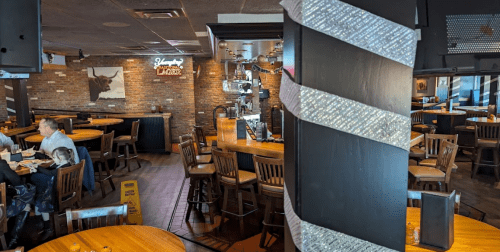 A cozy restaurant interior with wooden tables, a bar, and a decorative column, featuring a cow painting on the wall.