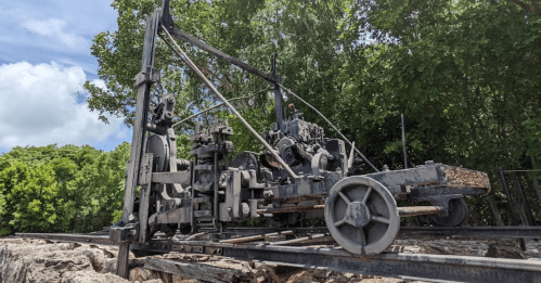 An old, rusted machine on tracks surrounded by greenery under a blue sky.