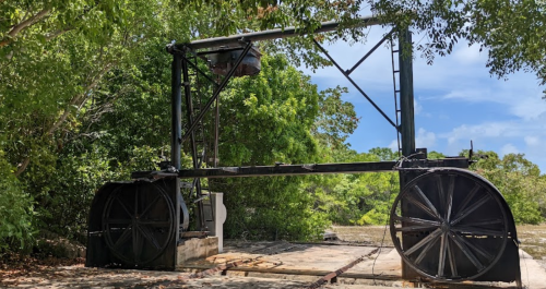 An old, large black structure with two big wheels, surrounded by lush greenery and a blue sky.