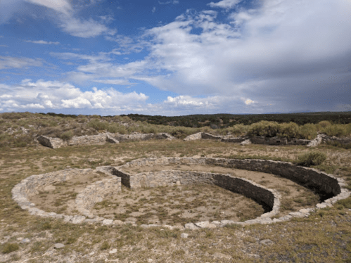 Ancient stone ruins in a circular formation, set against a cloudy sky and grassy landscape.