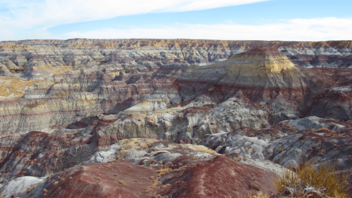 Vast, colorful badlands with layered rock formations under a blue sky, showcasing shades of red, brown, and gray.