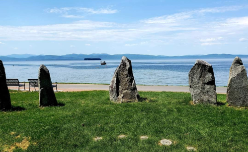 Five large stones stand on green grass by a calm lake, with distant mountains and a clear blue sky in the background.