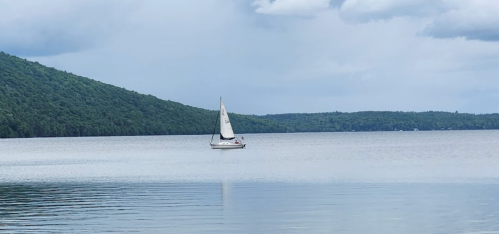 A sailboat glides across a calm lake, surrounded by lush green hills under a cloudy sky.