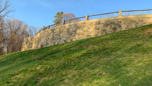 A stone wall with a wooden railing atop a grassy slope, surrounded by trees under a clear blue sky.