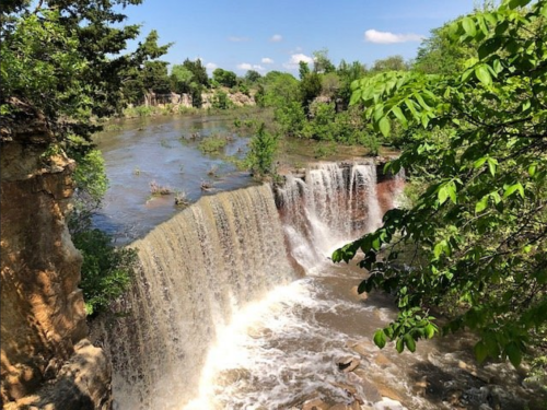 A scenic waterfall cascading over rocky cliffs, surrounded by lush greenery and a clear blue sky.
