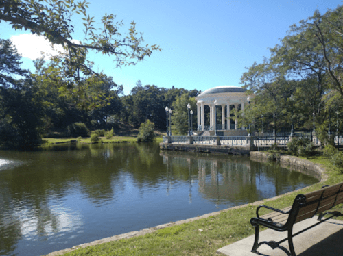 A serene park scene featuring a gazebo by a calm pond, surrounded by trees and a bench in the foreground.
