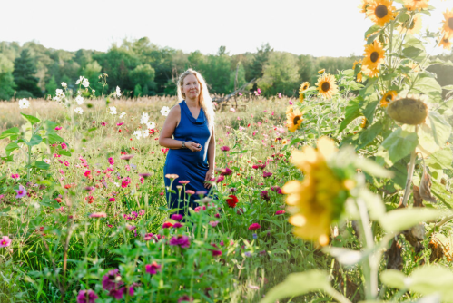 A woman in a blue dress stands among colorful flowers in a sunny field, surrounded by sunflowers and greenery.