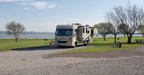 A parked RV by a lake, surrounded by grass and trees, with picnic tables in the background under a clear blue sky.