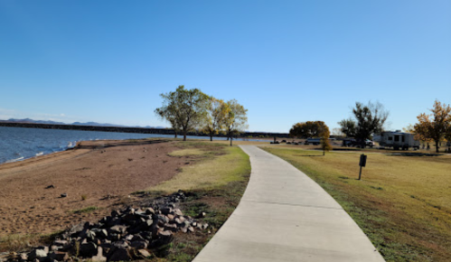 A scenic view of a pathway along a lake, with trees and a sandy shore under a clear blue sky.