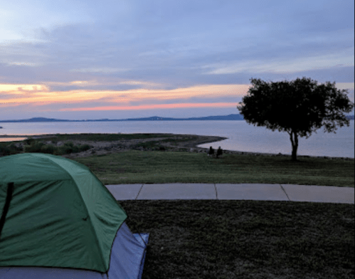 A green tent sits on grass near a lake at sunset, with a tree and distant hills visible in the background.
