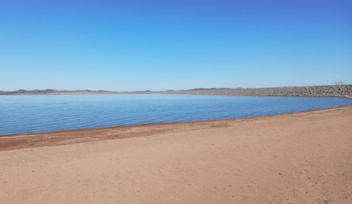 A calm lake with a sandy shore under a clear blue sky, surrounded by distant hills.