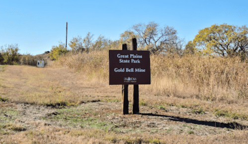 Sign for Great Plains State Park and Gold Bell Mine, surrounded by tall grass and trees under a clear blue sky.