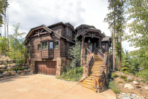A rustic wooden house with stone accents, featuring a large staircase and surrounded by trees and rocks.