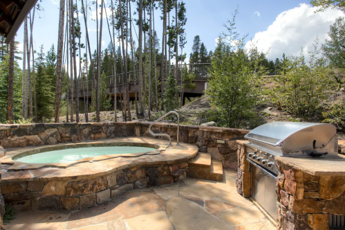 A stone patio features a hot tub and a grill, surrounded by trees and a clear blue sky.