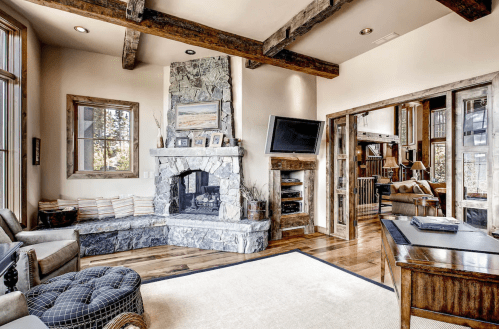 Cozy living room with stone fireplace, wooden beams, and large windows, featuring a comfortable seating area and a desk.