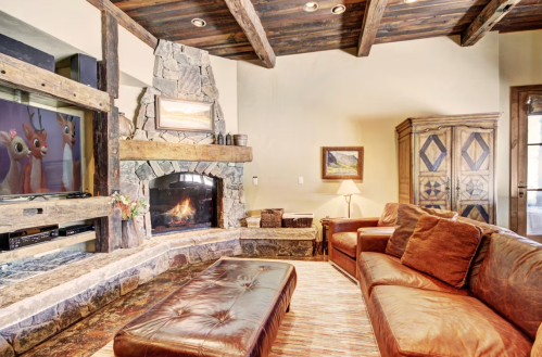 Cozy living room with a stone fireplace, wooden beams, a large sofa, and a TV featuring a deer-themed artwork.