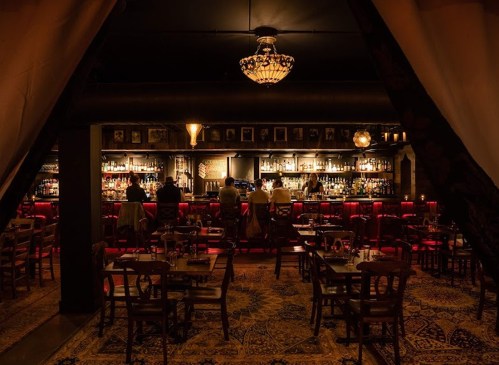 Dimly lit bar interior with a chandelier, wooden tables, and patrons at the bar, creating a cozy atmosphere.