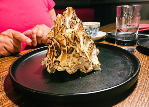 A large, toasted meringue dessert shaped like a mountain on a black plate, with a glass of water in the background.