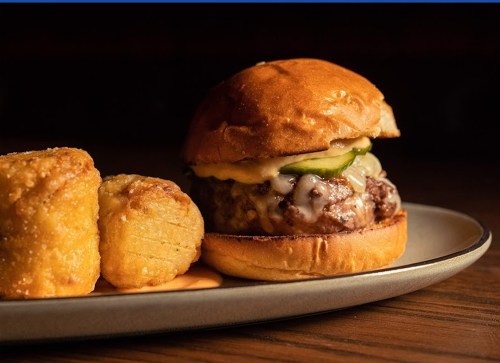 A juicy cheeseburger with pickles on a toasted bun, served with two golden, crispy potato bites on a plate.