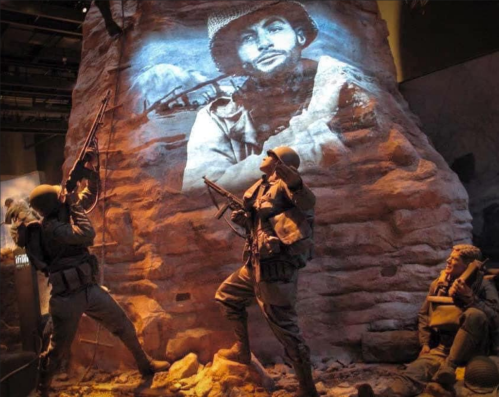 Soldiers in a museum exhibit, with a projected image of a soldier on a rocky wall behind them.