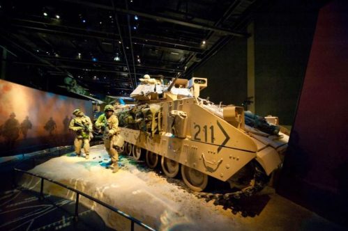 A military vehicle displayed in a museum, with soldiers in uniform nearby, set in a dimly lit environment.
