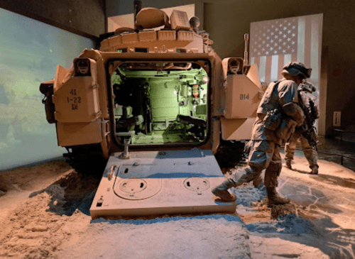 A military vehicle displayed in a museum, with soldiers interacting near it and an American flag in the background.