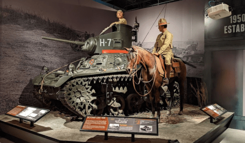 A tank displayed in a museum with a soldier on horseback beside it, featuring informational plaques in the foreground.