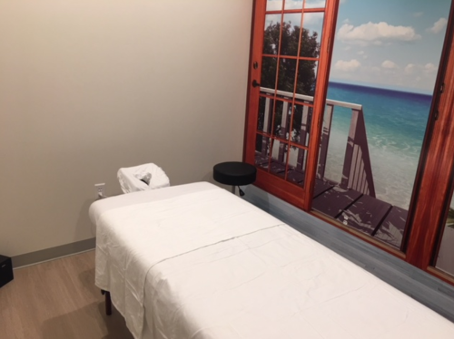 A treatment room with a massage table, a waste bin, and a large window mural of a beach scene.