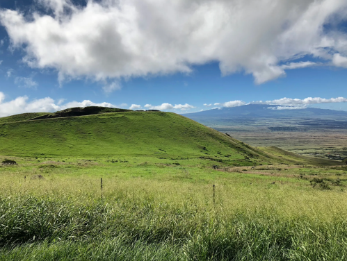 Lush green hills under a partly cloudy sky, with distant mountains and a vast landscape in the background.
